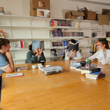Deux bibliothécaires discutent avec deux adolescentes, celles-ci ont le visage caché par un livre ouvert devant elles. 